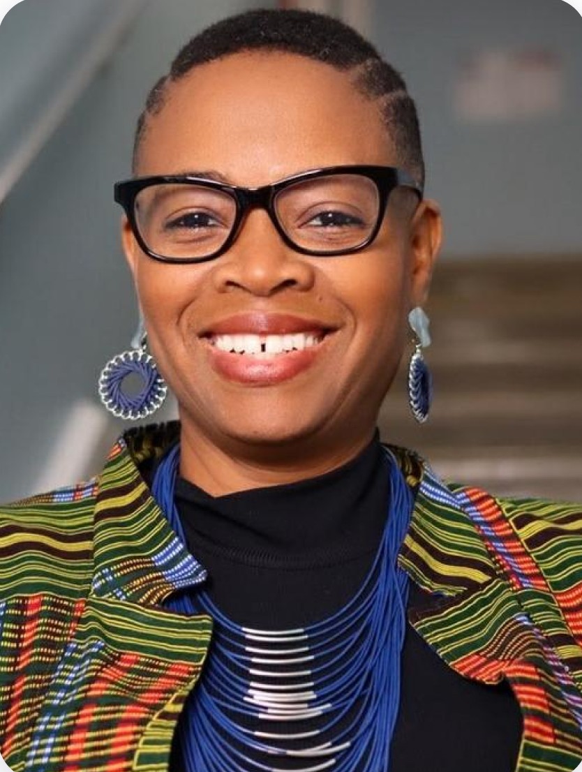 A Black woman with short hair, wearing a patterned blazer, black glasses, blue multi-stringed necklace and yarn-like loop earrings, smiles at the camera.